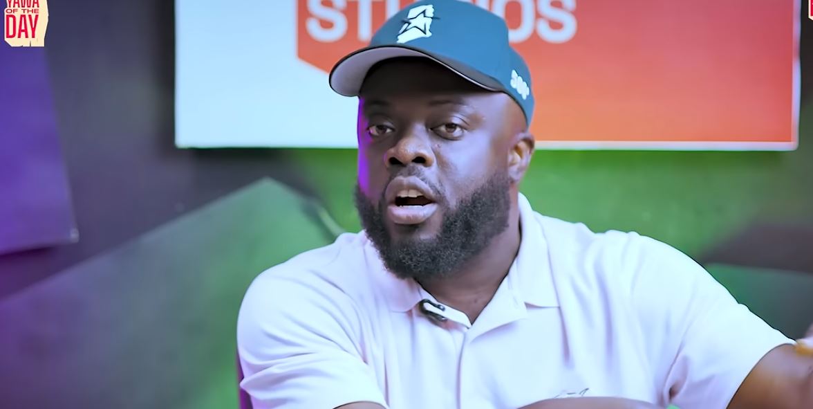 Man with beard in a light polo shirt and blue cap speaks on a colorful studio set with a red sign behind him